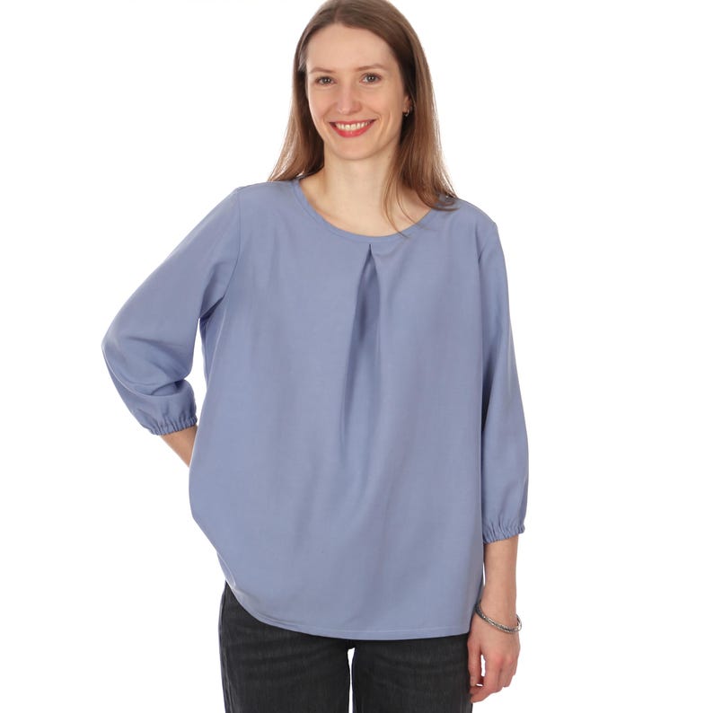 Pu&ograve; includere: Camicetta azzurra con scollo tondo e maniche a tre quarti. Le maniche sono raccolte ai polsini. La camicetta ha un dettaglio a pieghe sul davanti e un orlo curvo. La modella indossa jeans neri.
