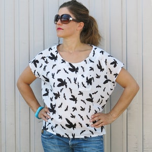 May include: A white blouse with a black bird print and a small pocket on the left side. The blouse has a scoop neck and short sleeves.