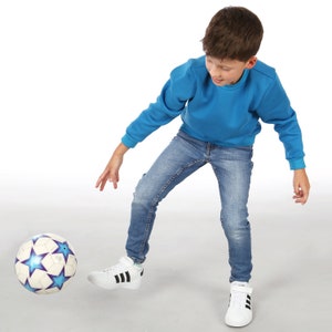 Könnte beinhalten: Ein junges Kind in blauen Jeans und einem blauen Sweatshirt tritt einen weißen Fußball mit blauen Sternen. Der Ball liegt auf einer weißen Oberfläche.