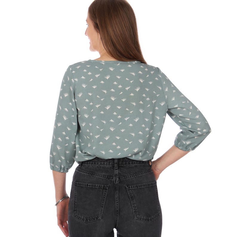 Pu&ograve; includere: Una camicetta verde chiaro con un motivo geometrico bianco. La camicetta ha maniche a tre quarti e scollo a V. La persona indossa jeans neri. La camicetta &egrave; realizzata in un tessuto morbido e fluido.