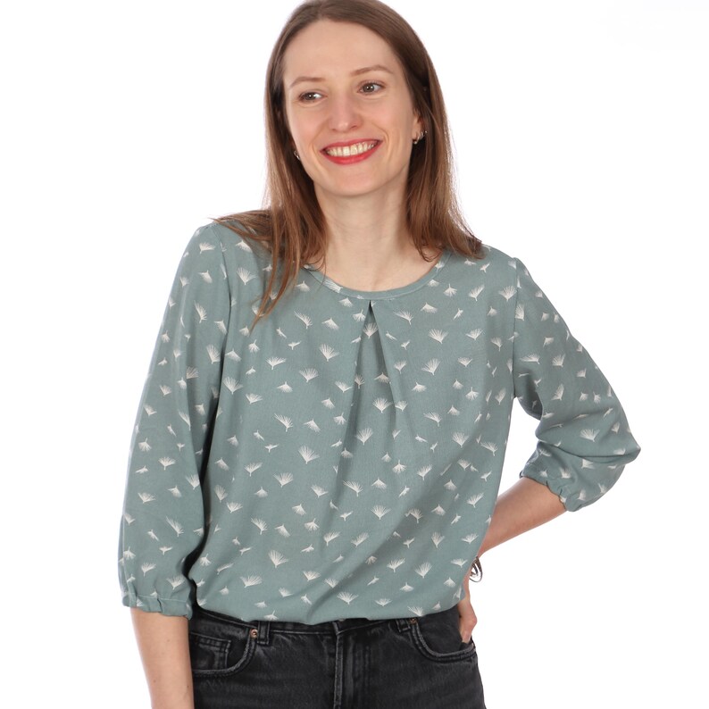 Pu&ograve; includere: Una camicetta verde acqua chiaro con scollo tondo e maniche a tre quarti. La camicetta presenta un delicato motivo bianco e un dettaglio a pieghe sullo scollo. Le maniche sono raccolte ai polsini. La modella indossa jeans neri.