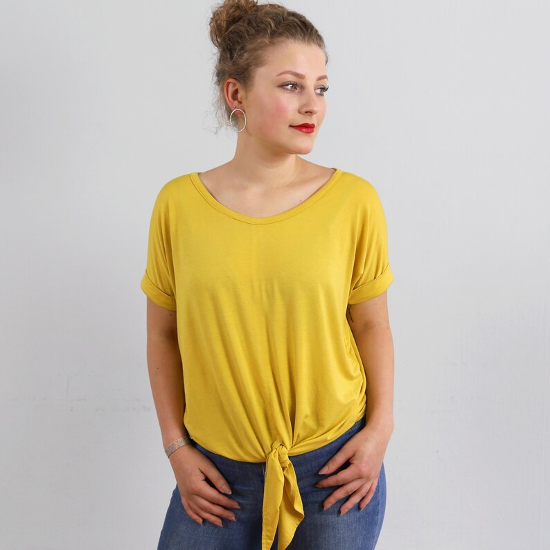 May include: A mustard yellow short-sleeved t-shirt featuring a round neckline, cuffed sleeves, and a knotted hem detail at the front. The shirt is paired with blue denim jeans, creating a casual look with a simple, solid-colored fabric design.