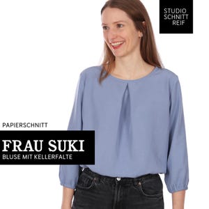 Pu&ograve; includere: Una camicetta azzurra con maniche a tre quarti e un davanti a pieghe. L'immagine include il testo "PAPIERSCHNITT FRAU SUKI BLUSE MIT KELLERFALTE" e "STUDIO SCHNITT REIF" in riquadri di testo in bianco e nero.