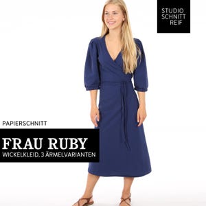 Può includere: Un abito blu navy avvolgente con scollo a V e maniche a 3/4. L'abito è legato in vita con una cintura abbinata. L'abito è etichettato "Frau Ruby Wickelkleid, 3 Ärmelvarianten".