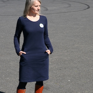 Op de afbeelding: Marineblauwe jurk met lange mouwen, ronde hals en zakken. De jurk heeft een knielange zoom en een kleine ronde speld. De persoon draagt bruine laarzen.