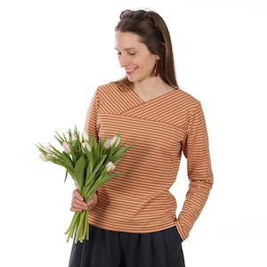 Puede incluir: Una blusa de manga larga con diseño cruzado a rayas horizontales en tonos marrón y crema. La persona lleva pantalones oscuros y sostiene un ramo de tulipanes blancos y rosas. Gafas de sol en la cabeza.