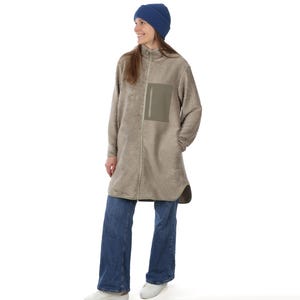 Puede incluir: Una mujer con un gorro azul, una chaqueta larga de forro polar beige con un bolsillo en el pecho, vaqueros azules y zapatillas blancas. La chaqueta tiene una cremallera completa y un cuello alto. El atuendo es informal y cómodo.