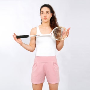 Könnte beinhalten: Eine Frau in einem weißen Tanktop und rosa Shorts hält einen weißen und braunen Squashschläger.