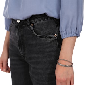 Pu&ograve; includere: Primo piano di una persona che indossa una camicetta azzurra e jeans neri. La persona indossa anche un braccialetto d'argento. I jeans hanno un classico design a cinque tasche.