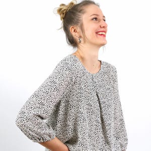 Pu&ograve; includere: Una camicetta bianca con un motivo a pois neri. La camicetta ha maniche lunghe e scollo tondo. La donna sorride e guarda in alto. Le maniche sono arricciate ai polsini.