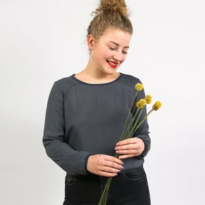 Könnte beinhalten: Eine Frau trägt ein dunkelgraues Langarmshirt mit Rundhalsausschnitt und schwarze Hose und hält einen Strauß getrockneter gelber Blumen.