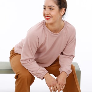 Peut inclure: Une femme portant un sweat-shirt rose poudré à manches longues et un détail plissé aux poignets. Elle porte un pantalon marron et est assise sur un banc vert clair.