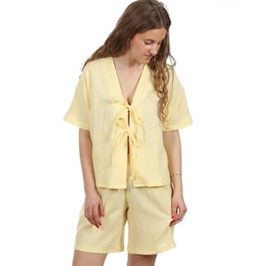 May include: A light yellow two-piece pajama set. The top has short sleeves and a V-neck with tie closures. The matching shorts are knee-length. The fabric has a textured appearance.