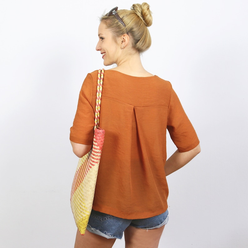 May include: A woman wearing a rust-colored, short-sleeved blouse with a back pleat. She is carrying a multi-colored woven tote bag.