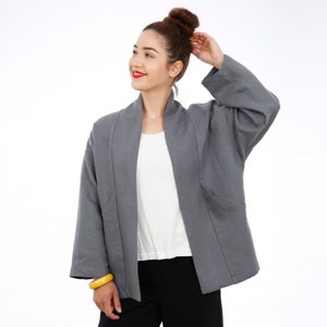 Peut inclure: Une veste en lin gris avec un devant long et ouvert et une coupe d&eacute;contract&eacute;e. La veste a un col et des manches longues. Le mannequin porte la veste sur un t-shirt blanc et un pantalon noir.
