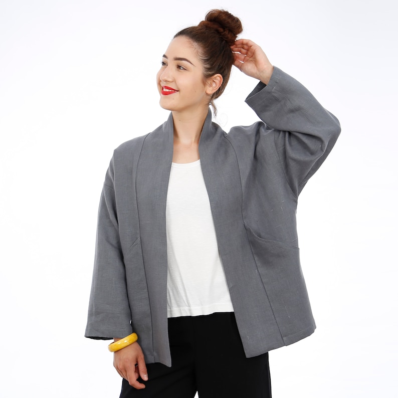 Peut inclure: Une veste en lin gris avec un devant long et ouvert et une coupe d&eacute;contract&eacute;e. La veste a un col et des manches longues. Le mannequin porte la veste sur un t-shirt blanc et un pantalon noir.