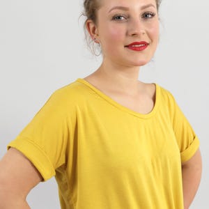 May include: A mustard yellow t-shirt with a wide scoop neckline and short sleeves featuring folded cuffs. The garment is made of a solid-colored, lightweight fabric that drapes loosely over the torso, providing a simple and clean aesthetic.