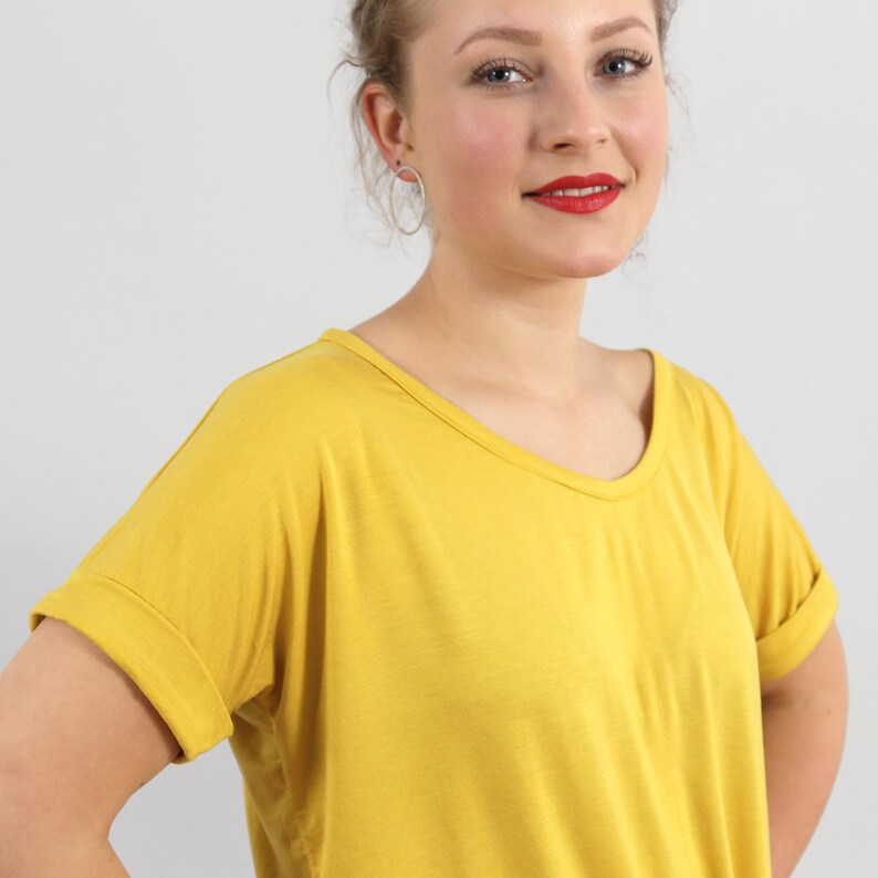 May include: A mustard yellow t-shirt with a wide scoop neckline and short sleeves featuring folded cuffs. The garment is made of a solid-colored, lightweight fabric that drapes loosely over the torso, providing a simple and clean aesthetic.