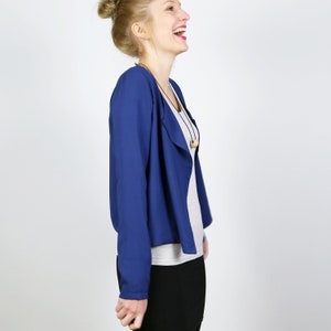 Pode incluir: Uma mulher vestindo um blazer azul aberto na frente com uma camisa cinza por baixo. Ela est&aacute; rindo e olhando para a direita.