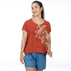 May include: A woman wearing a rust-colored, short-sleeved, loose-fitting top with a round neckline and blue denim shorts. She is holding a bouquet of dried white flowers.