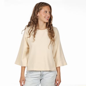 Könnte beinhalten: Ein cremefarbenes Langarmshirt mit Rundhalsausschnitt und weiten, ausgestellten Ärmeln. Das Kleidungsstück hat einen geraden Saum und besteht aus einem einfarbigen, neutralen Stoff, kombiniert mit einer hellblauen Jeans mit sichtbarem Knopf.