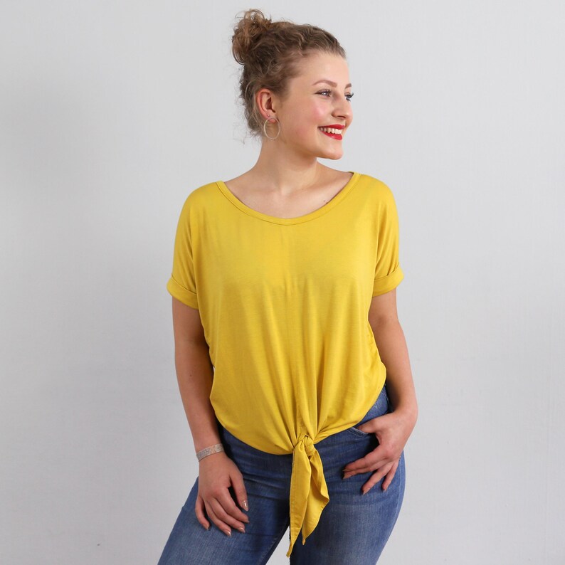 May include: A mustard yellow short-sleeved t-shirt featuring a wide scoop neckline and a knotted front hem detail. The shirt is paired with blue denim jeans, creating a casual look with rolled sleeve cuffs and a relaxed fit throughout the torso.