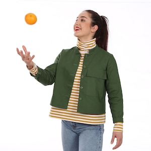 Könnte beinhalten: Eine Frau trägt eine grüne Jacke über einem gestreiften Shirt und einer blauen Jeans und wirft eine Orange in die Luft.