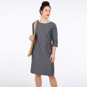 Op de afbeelding: Een grijze, knielange jurk met een ronde hals en driekwart mouwen. De jurk heeft een bruine leren riem die in de taille is geknoopt. Het model draagt de jurk en houdt een geweven strooien tas.