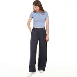 Puede incluir: Una mujer con una camiseta azul claro de manga corta y pantalones anchos azul marino oscuro. Los pantalones tienen cintura alta y bolsillos. Lleva zapatillas blancas. El fondo es blanco liso.