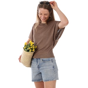 May include: A woman wearing a brown short-sleeved top and denim shorts, holding a woven basket filled with yellow tulips. Sunglasses are perched on her head. The background is plain white.