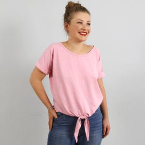 May include: A light pink short-sleeved top with a round neckline and a center vertical seam. The garment features cuffed sleeves and a fabric tie detail at the front hem, creating a gathered effect. The material has a soft, textured appearance.