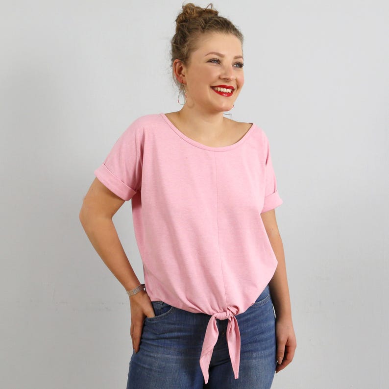 May include: A light pink short-sleeved top with a round neckline and a center vertical seam. The garment features cuffed sleeves and a fabric tie detail at the front hem, creating a gathered effect. The material has a soft, textured appearance.