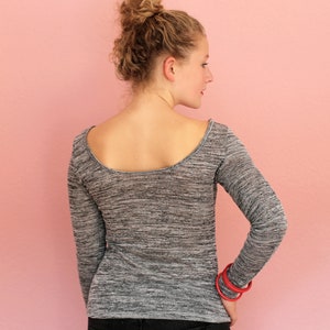 May include: A gray long-sleeve shirt with a scoop back. The shirt has a textured pattern and is worn with black pants. The person is wearing red bracelets. The background is a solid pink color.