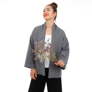 Könnte beinhalten: Eine anthrazitfarbene, vorne offene Jacke mit weiten Ärmeln und Schalkragen, getragen über einem weißen Oberteil und einer schwarzen Hose, während ein Strauß aus getrockneten violetten und weißen Blumen vor weißem Hintergrund gehalten wird.