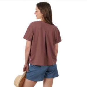 Könnte beinhalten: Ein kurzärmliges, malvenfarbenes Oberteil mit einem plissierten Rückendetail. Kombiniert mit jeansblauen Shorts. Ein Strohhut wird in der Hand gehalten. Das Outfit ist lässig und für warmes Wetter geeignet.