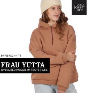 Könnte beinhalten: Eine Frau trägt einen übergroßen Kapuzenpullover in Tan mit einem Viertel-Reißverschluss. Der Hoodie hat eine Kapuze und lange Ärmel. Der Text "FRAU YUTTA" und "OVERSIZED HOODIE IM TROYER-STIL" werden in einem schwarzen Rechteck angezeigt. Eine cremefarbene Mütze wird getragen.