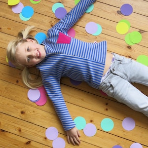 Op de afbeelding: Een jong persoon draagt een lang mouw shirt met blauwe en witte strepen met een roze hart op de voorkant, een blauwe spijkerbroek en vlechtjes. De persoon ligt op een houten vloer met kleurrijke papieren cirkels die eromheen verspreid zijn.