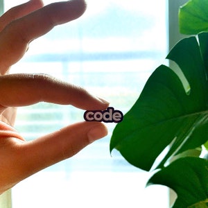 Puede incluir: Primer plano de un dedo sosteniendo un pin de esmalte negro y blanco con la palabra "código" escrita en letras blancas.