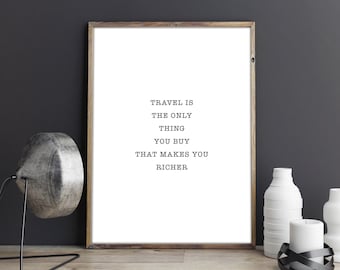 May include: A black and white typography print with the quote "Travel is the only thing you buy that makes you richer". The print is framed in a light brown wood frame and is displayed on a wooden surface with a black lamp and white vases.