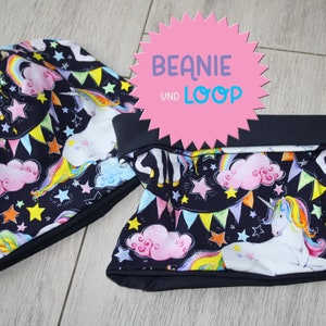 Może przedstawiać: Czarna czapka i komin z wzorem jednorożca, tęczy i gwiazd. Na różowej metce widnieje napis "BEANIE UND LOOP".
