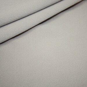 Könnte beinhalten: Nahaufnahme eines hellgrauen Stoffes mit einer weichen, strukturierten Oberfläche. Der Stoff ist über sich selbst gefaltet, wodurch Schatten entstehen und die Textur des Materials hervorgehoben wird. Das Gesamtbild deutet auf ein gemütliches, warmes Textil hin.