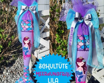 May include: A purple and blue mermaid-themed school cone with a silver ribbon and a tulle bow. The cone has a mermaid illustration and the text "Schulkind" and "Lena". The cone is decorated with a mermaid tail and scales. The text "Schultüte Meerjungfrau Lila" is on a blue starburst.