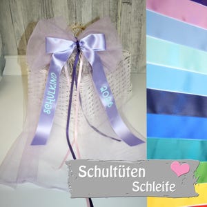 Può includere: Un fiocco decorativo con nastri color lavanda e la scritta "SCHULKIND 2026" è attaccato a un cestino di vimini bianco. Il fiocco è realizzato in raso e tessuto trasparente. Una tabella colori di nastri è a destra.