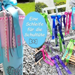 Op de afbeelding: Een blauwe en roze schoolkegel met een witte strik en de tekst "SCHULKIND" en "2023". De kegel is versierd met een witte kantversiering en een roze en wit stippenpatroon. De kegel is omringd door andere schoolkegels in verschillende kleuren en ontwerpen.