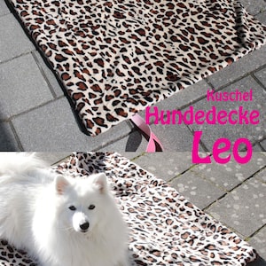 Könnte beinhalten: Ein rechteckiges Haustierbett mit Leopardenmuster in Braun- und Cremetönen. Das Bett hat ein rosa Band. Der Text "Leo" ist in Pink zu sehen. Ein weißer Hund liegt auf dem Bett.
