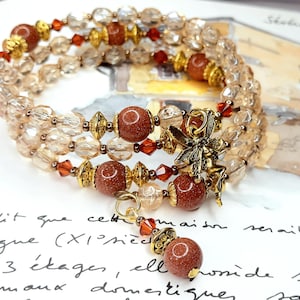 Könnte beinhalten: Ein dreireihiges Armband mit braunen und goldenen Perlen, einem goldenen Feen-Anhänger und einem braunen Stein-Anhänger.