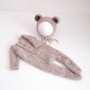 May include: A beige knitted baby outfit with a matching bear hat. The outfit features a long-sleeved jumpsuit with a button closure and a hood. The hat has bear ears and a tie.