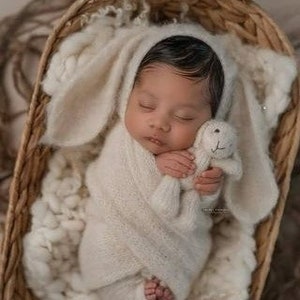 Könnte beinhalten: Ein neugeborenes Baby ist in eine weiße gestrickte Decke mit Hasenohren gewickelt. Das Baby hält ein weißes gestricktes Hasenspielzeug. Das Baby liegt in einem Weidenkorb.