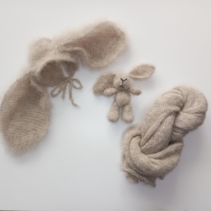 Könnte beinhalten: Ein Set aus drei beigefarbenen Strickartikeln für Neugeborenen-Fotografie-Requisiten. Das Set beinhaltet eine Hasenmütze, ein Hasenspielzeug und eine Wickel.