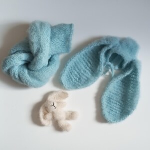 May include: A light blue knitted baby wrap and bunny ears set, along with a small cream-colored bunny toy. The set is made of soft, fluffy material, perfect for newborn photography or baby gifts. The bunny toy has a simple embroidered face.